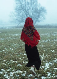 Figure in a red patterned headscarf and dark skirt running across a snowy field toward a bare tree in fog.