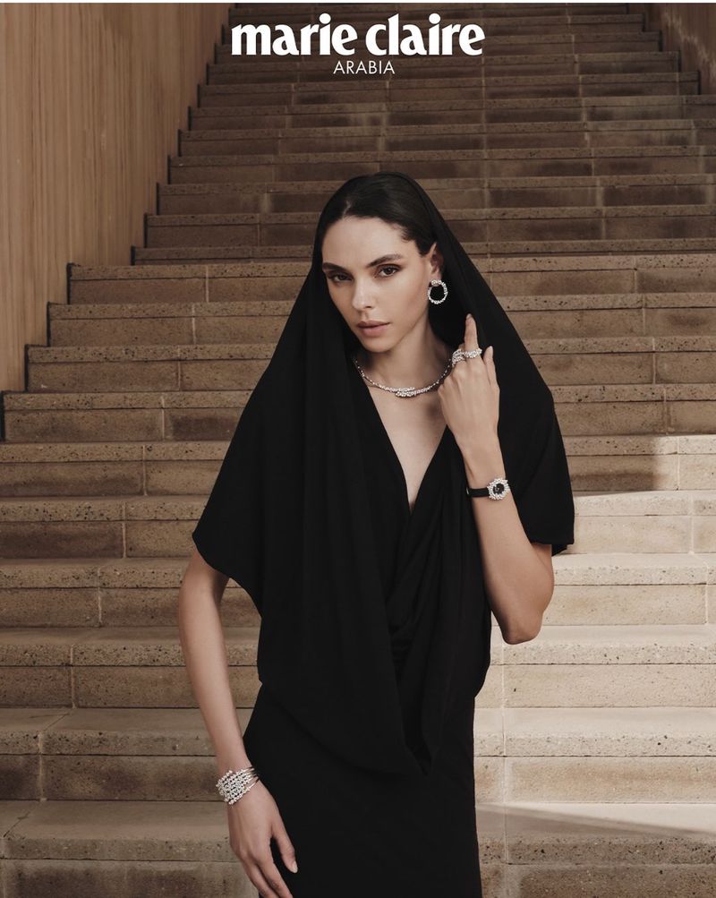 Model Aline Novais in a black dress with a draped head covering wearing diamond jewelry on stone stairs for Marie Claire Arabia