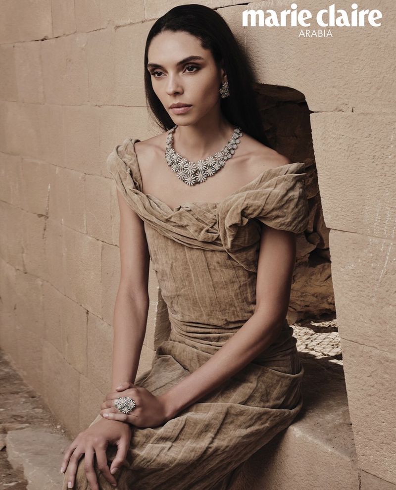 Brunette woman in tan dress with silver statement jewelry, posing in front of a tan stone wall for a Marie Claire Arabia fashion editorial.