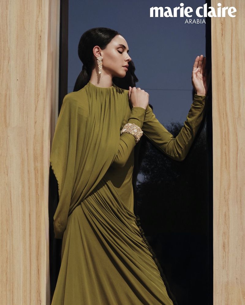 Model in olive green dress wearing ornate diamond earrings and cuff bracelet leaning against a wooden doorframe