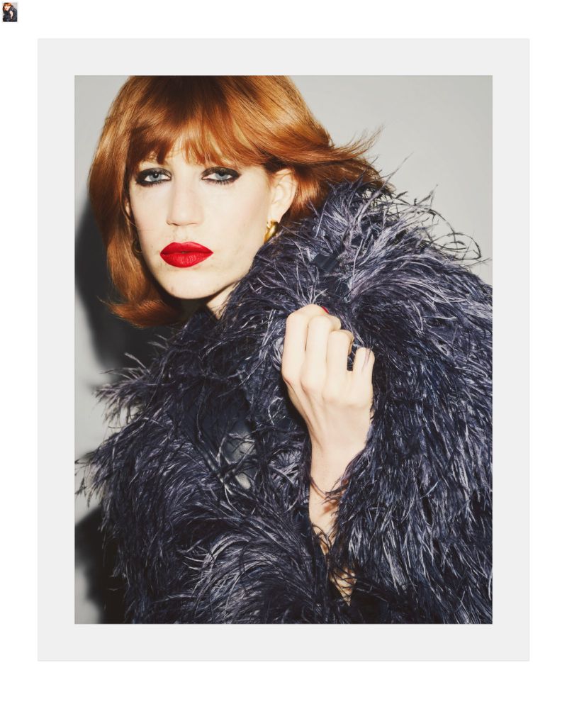 Close-up portrait of a red-haired female adult wearing a feathered jacket, red lipstick, and gold earrings against a grey background.