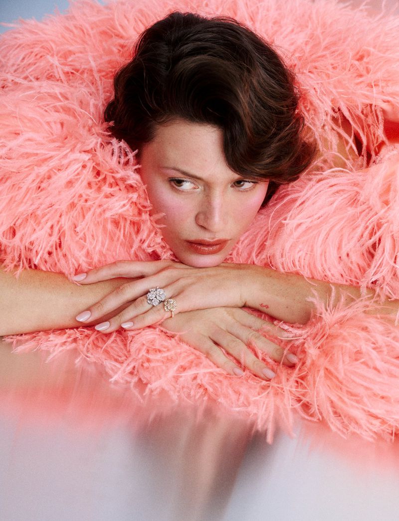 Model with dark hair wearing a fluffy pink feather boa and diamond rings, resting chin on hands against a pale background
