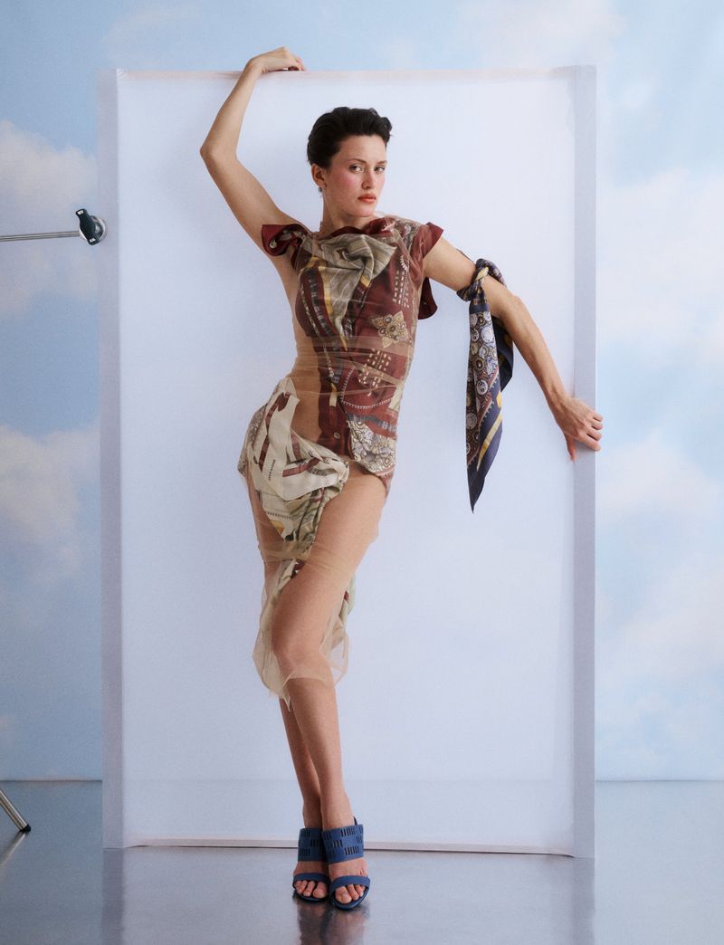 Model posing against a white backdrop with a printed dress and a silk scarf tied to her arm for Vogue Greece