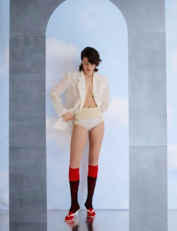 Model wearing a white blazer, pale underwear, and red socks standing beneath an architectural archway for Vogue Greece
