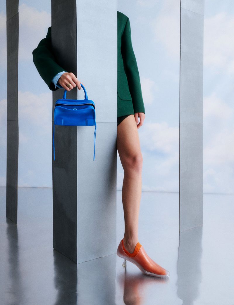 Fashion shot featuring a person in a green suit holding a blue purse, with orange shoes, against a reflective and architectural backdrop.