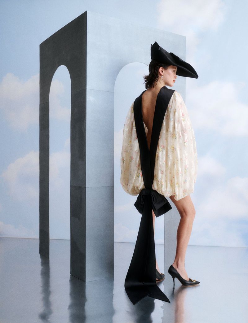 Model wearing a white dress with black bow details and black hat posing next to a grey arch structure with a cloudy sky background.