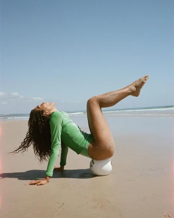 Woman in a green long-sleeve swimsuit balancing on a volleyball on a sandy beach under a clear sky.