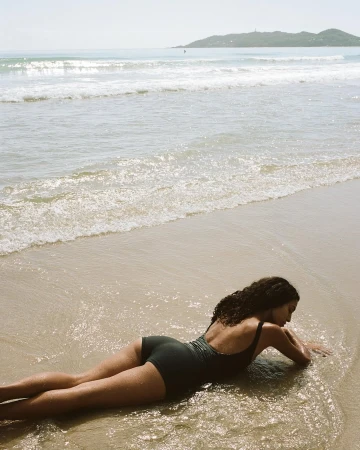 Woman in a one-piece swimsuit lies on a sandy beach at the ocean's edge, with gentle waves washing around her and a distant island.