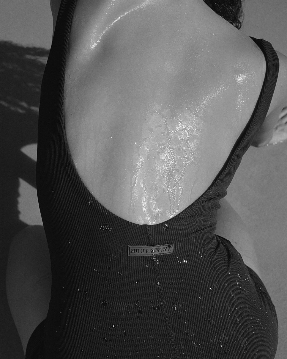 Black and white close-up photo of an adult female wearing a black swimsuit, seen from the back, with water droplets on her skin.