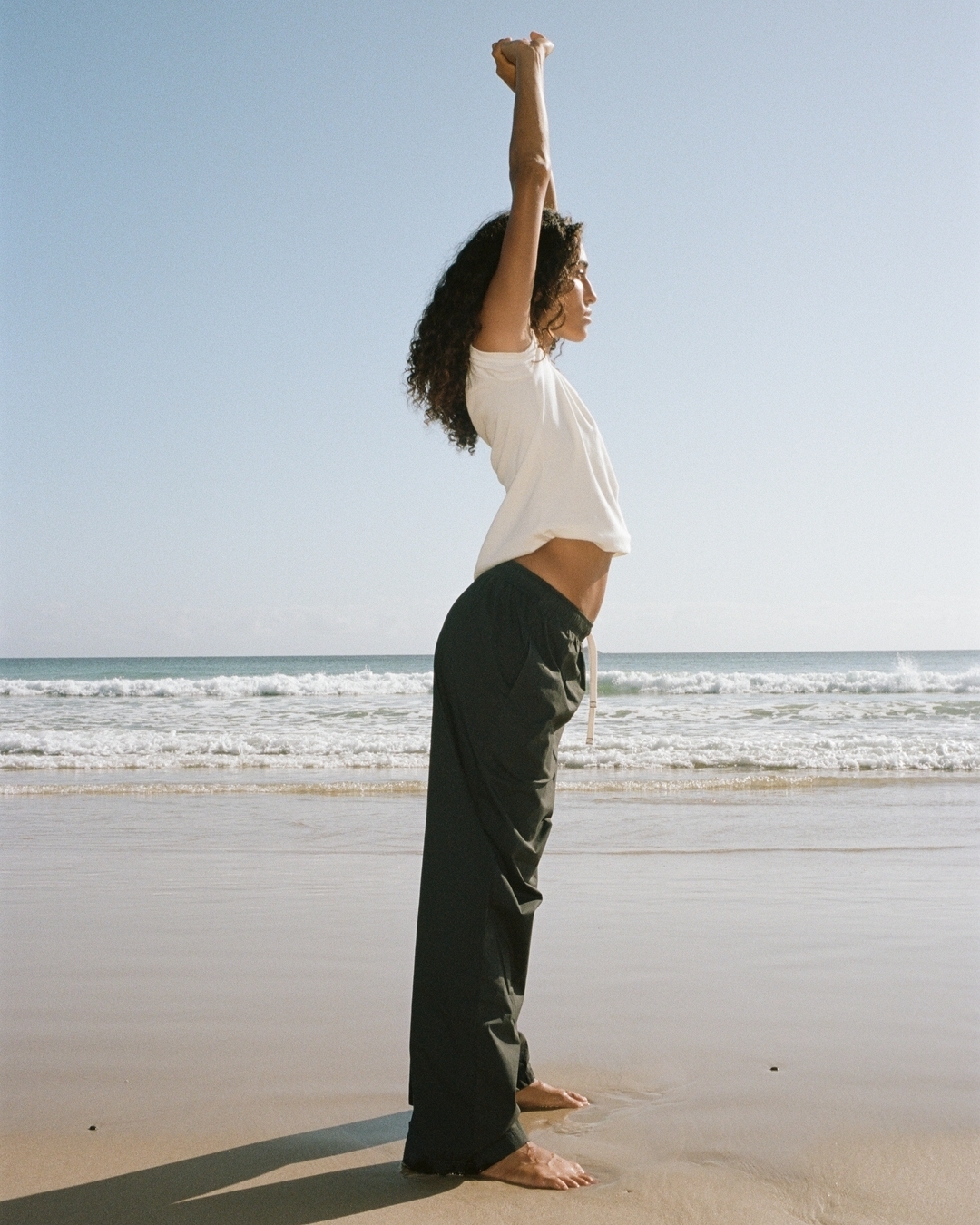 Profile view of a woman with long dark curly hair wearing a white top and dark trousers stretching arms upward on a sandy beach.