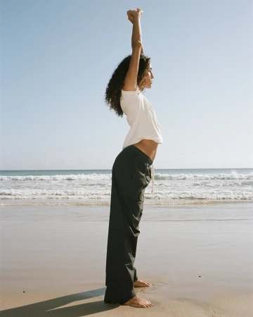 Profile view of a woman with long dark curly hair wearing a white top and dark trousers stretching arms upward on a sandy beach.