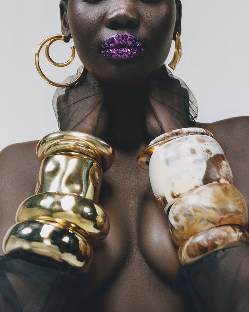 Close-up of a Black woman with purple glitter lipstick wearing chunky gold earrings and bracelets against a white background.