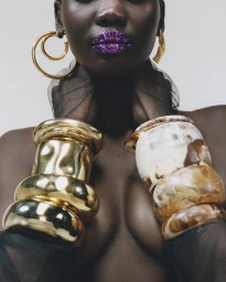 Model Achol Ayor wearing sparkling purple lipstick, chunky gold cuffs, and marbled bangles against a neutral studio backdrop.