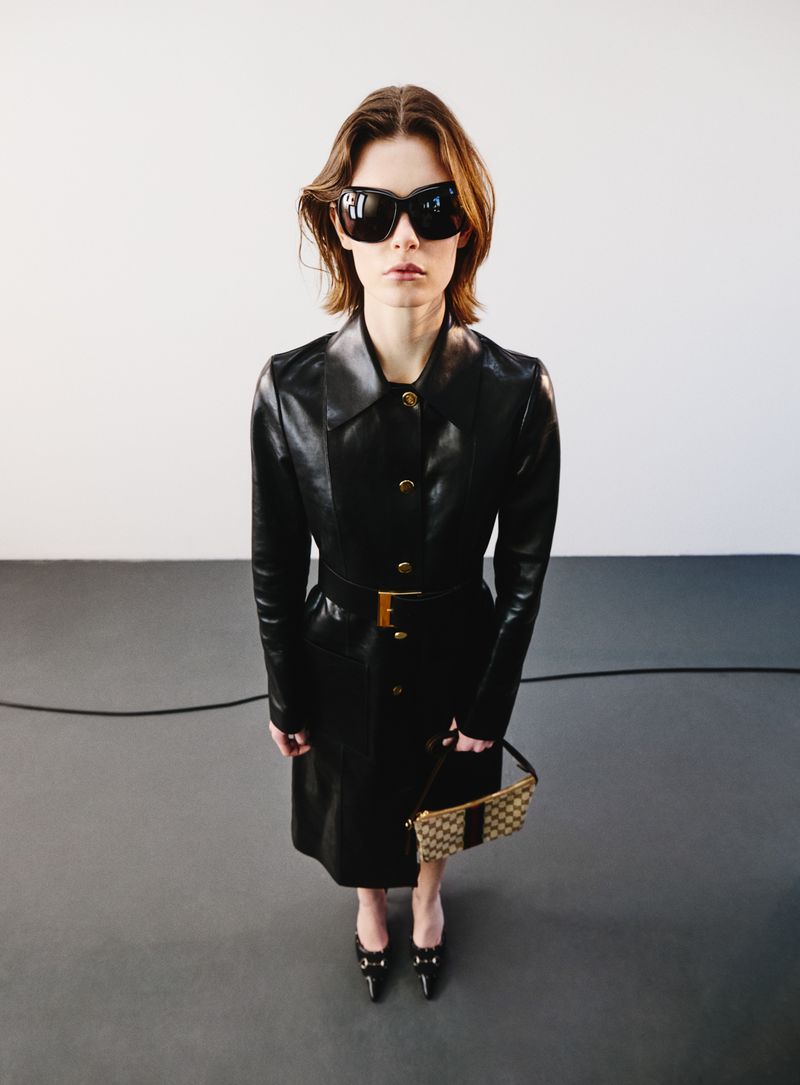 Model in oversized sunglasses wearing a black leather coat holding a small handbag against a studio backdrop