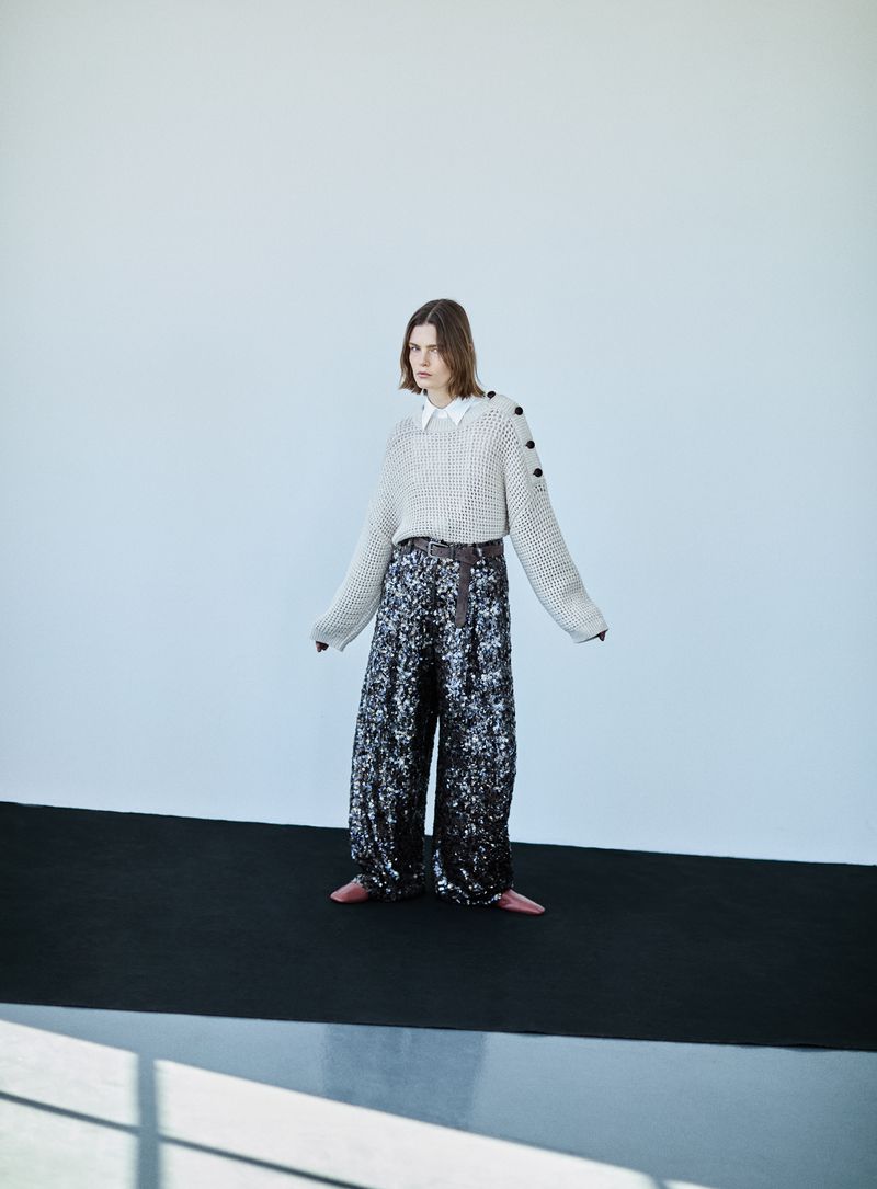 Model standing on a black surface against a white wall wearing a knitted sweater and sequin trousers