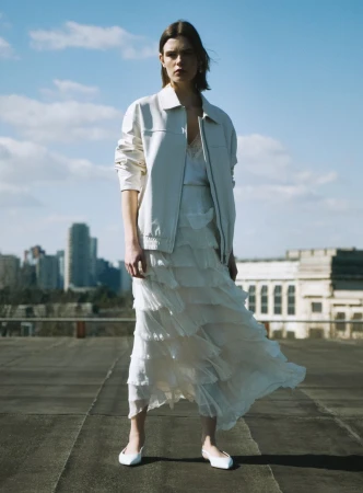 Model in white layered skirt and jacket standing on a rooftop with city skyline