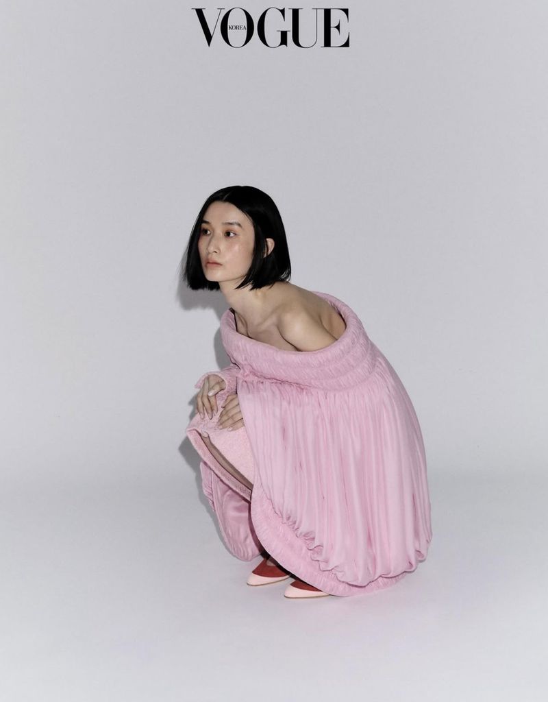Model Esther Kim in a pink off-the-shoulder dress crouching against a plain light grey studio background for Vogue Korea editorial