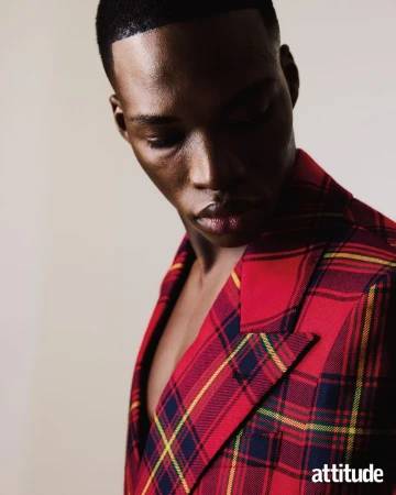 Portrait of a Black man looking down wearing a red plaid blazer against a plain neutral background for Attitude Magazine