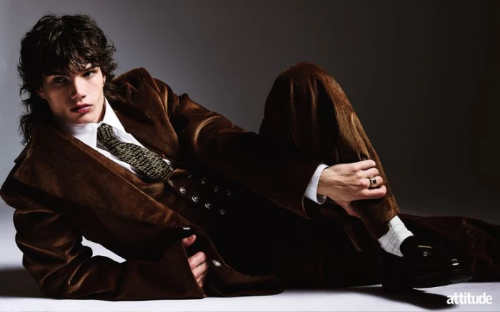 A male model reclining on a studio floor wearing a brown corduroy suit, white shirt, patterned tie, and loafers.