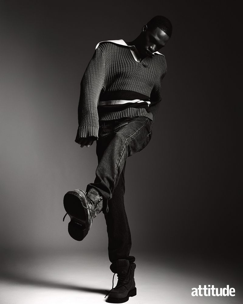 Monochrome full shot of a black male model in jeans and a striped sweater stepping forward, lit from above on a graduated gray background.