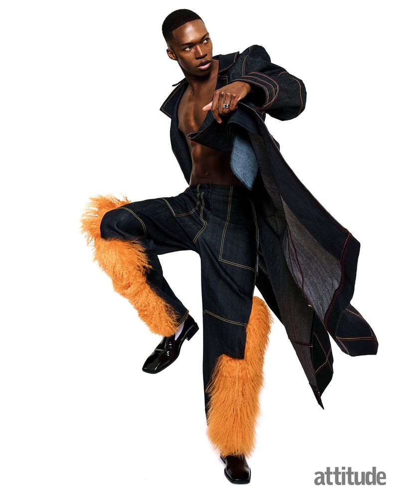 Black man wearing dark denim trench coat and feathered trousers posing against a bright white background