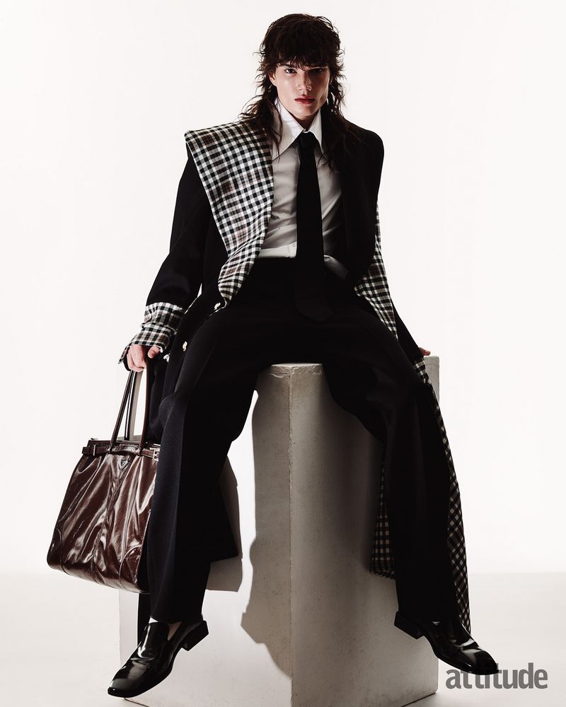 Model with long dark hair seated on a grey block wearing a black suit jacket with houndstooth lapel and holding a brown patent tote bag.