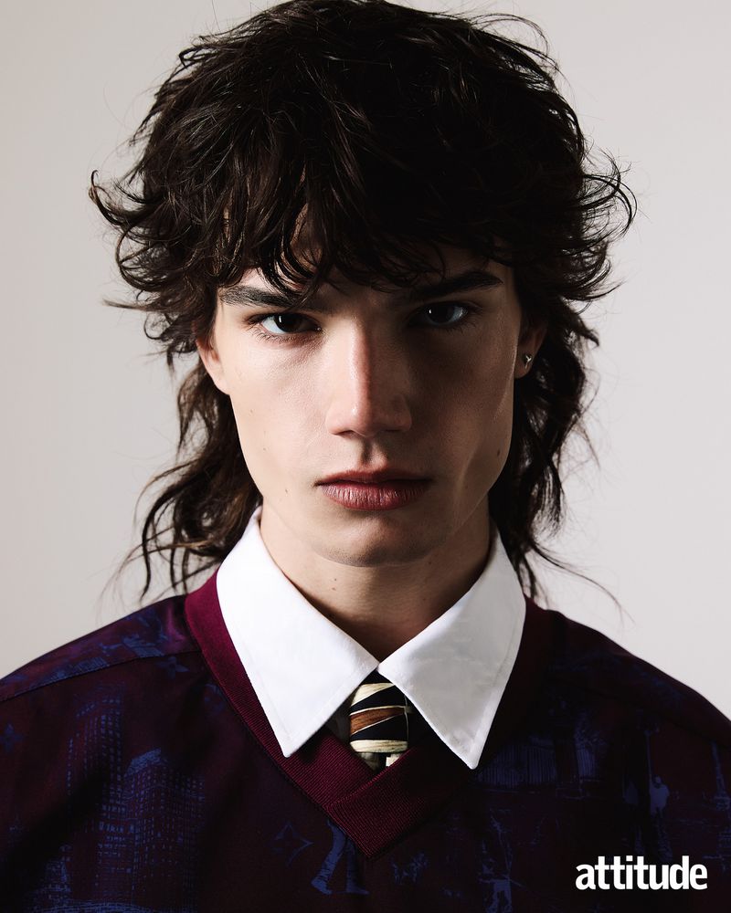 Portrait of a young man with dark tousled hair wearing a burgundy patterned sweater, white collared shirt, and striped tie.