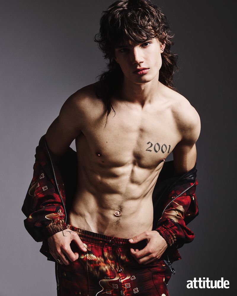 Shirtless young man with dark wavy hair and a tattoo on his chest wearing red patterned trousers against a grey backdrop