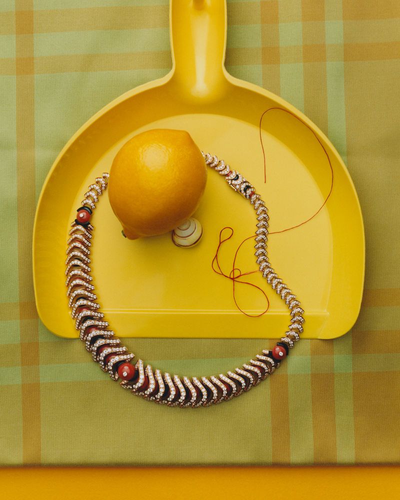 A yellow dustpan holds a whole lemon and an ornate jeweled necklace on a green patterned background for American Vogue.