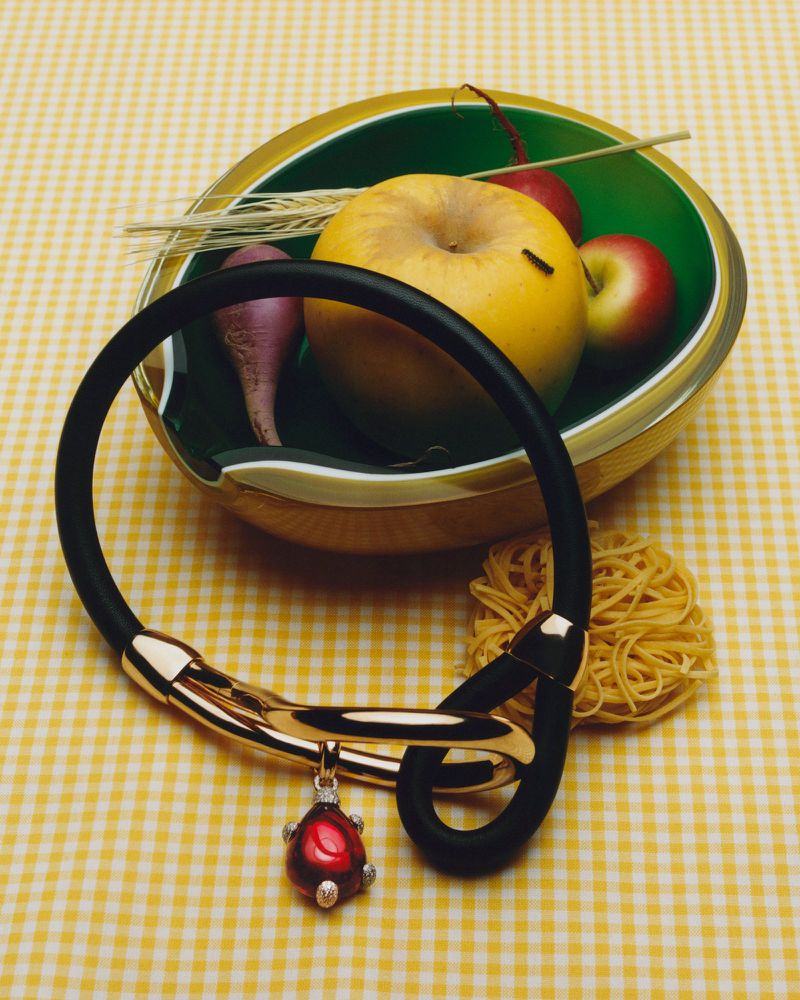 Close-up still life editorial image featuring a gold and black necklace with a ruby pendant resting on a yellow gingham background beside a fruit bowl.