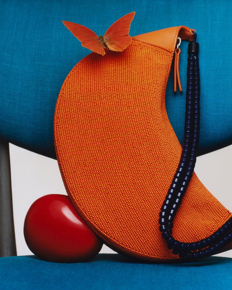 Still life composition featuring an orange beaded purse adorned with an orange butterfly, a red sphere, set against a blue fabric backdrop.