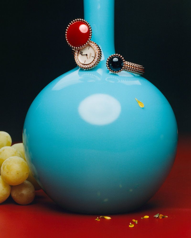 Close-up product photography of luxury rings and a wristwatch resting on a bright turquoise vase with pale grapes on a red surface