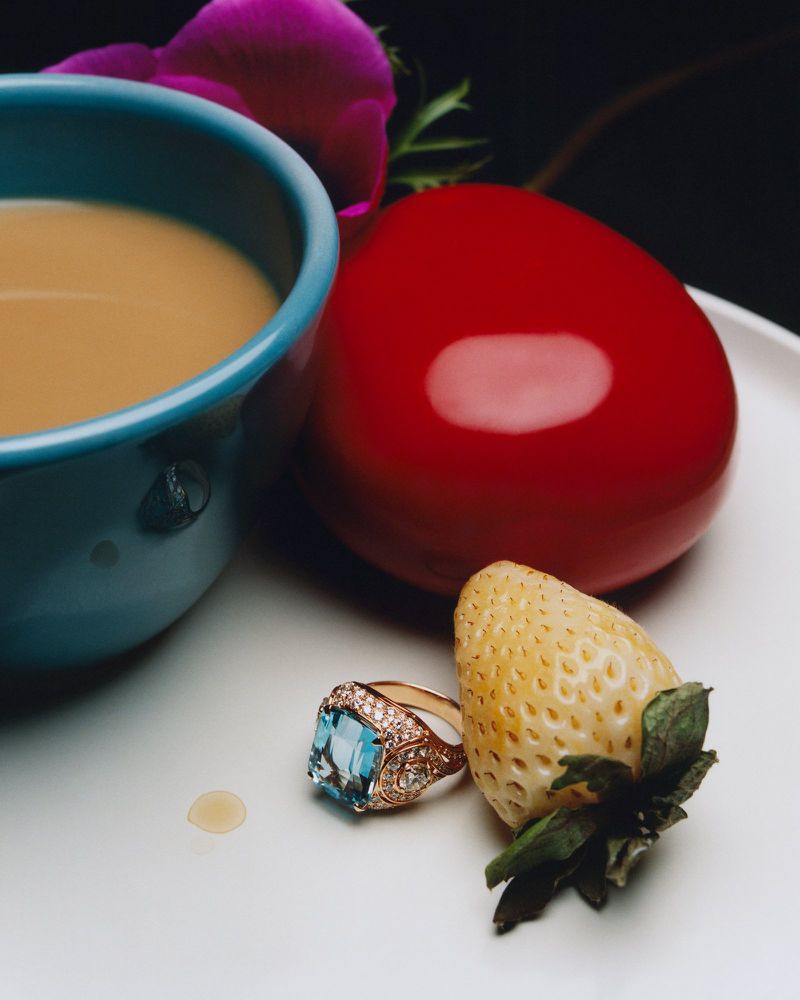 Close-up still life with a blue ceramic cup, red glossy ornament, white strawberry, and an ornate gemstone ring on a white plate.