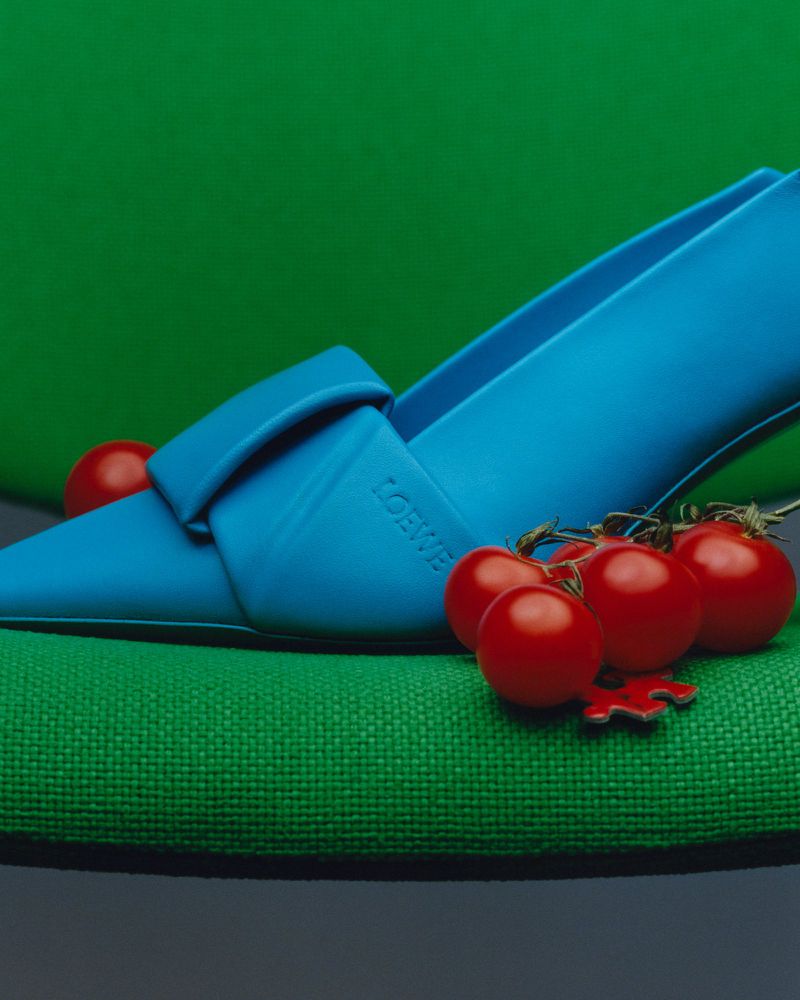 Bright blue pointed-toe shoe resting on a textured green chair beside a cluster of small red tomatoes for American Vogue Color Guard.