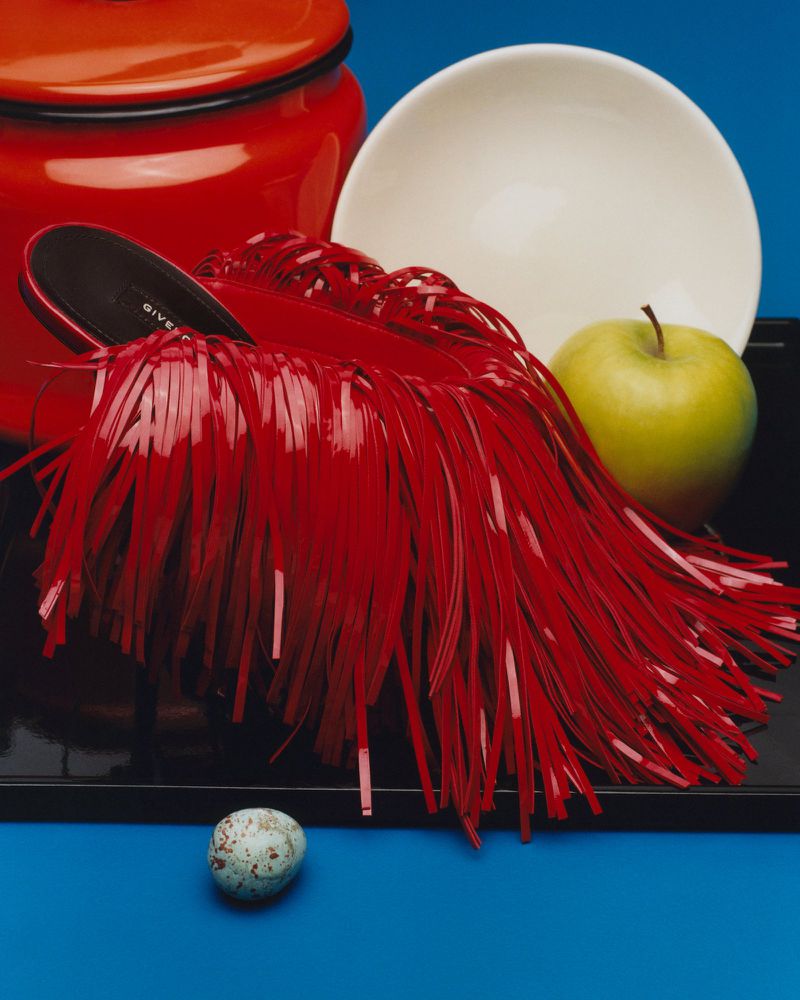 Red fringed mule shoe resting on a tray with a green apple, white bowl, and red canister against a vivid blue background.
