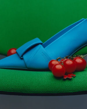 Bright blue pointed-toe shoe resting on a textured green chair beside a cluster of small red tomatoes for American Vogue Color Guard.