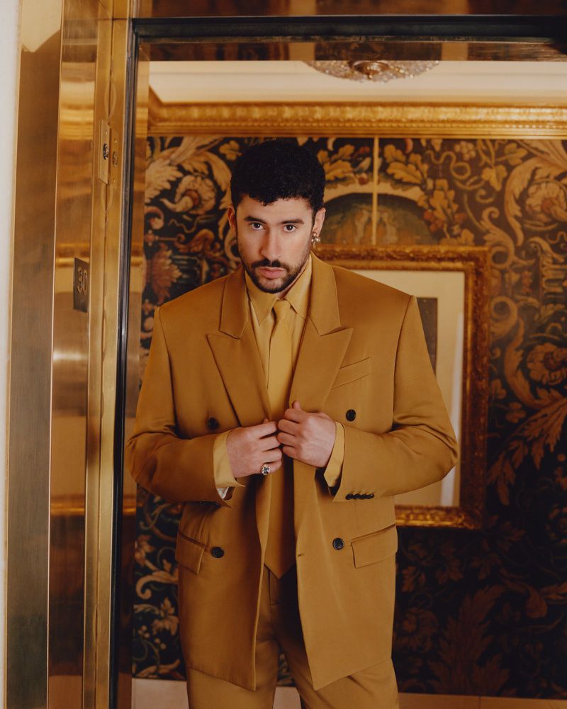Bad Bunny wearing a tailored camel suit in an elevator with patterned wallpaper