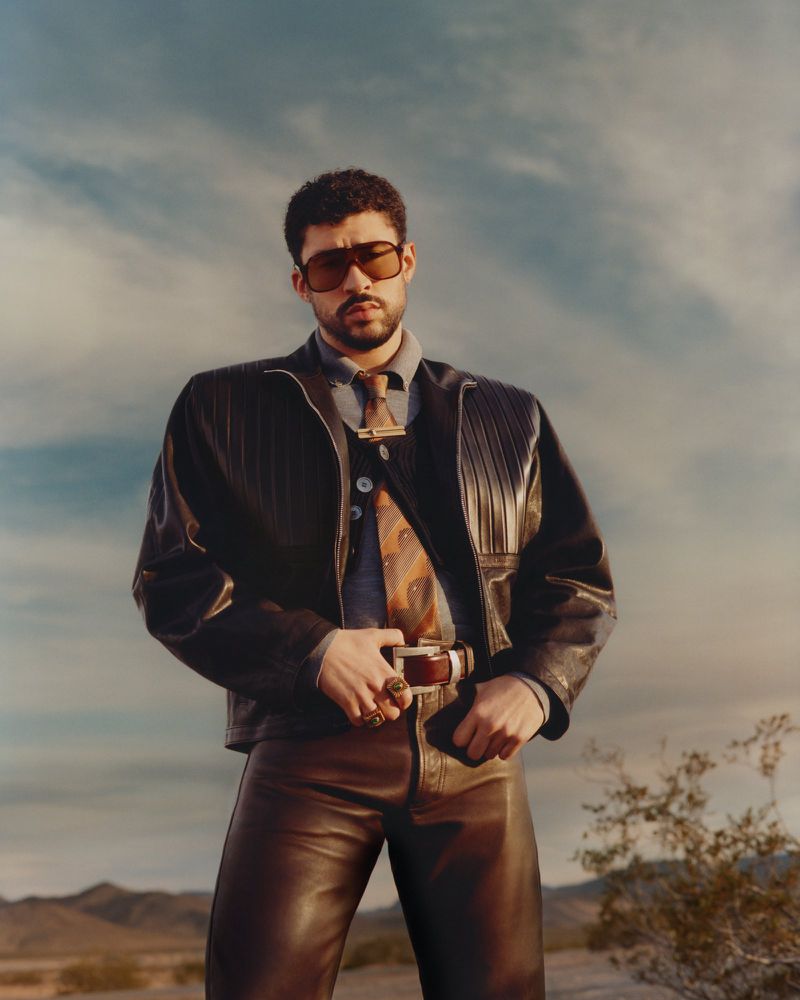 Adult man wearing sunglasses, leather jacket, pants and tie poses in a desert landscape with mountains and blue sky in the background.