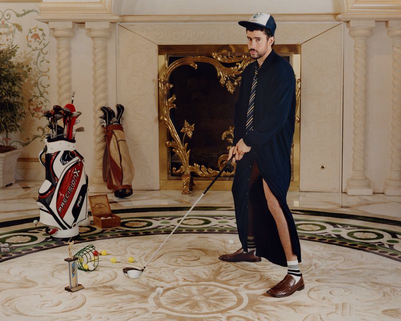 Bad Bunny posing with a golf club in a luxury hotel lobby wearing a long dark coat and cap for American Vogue