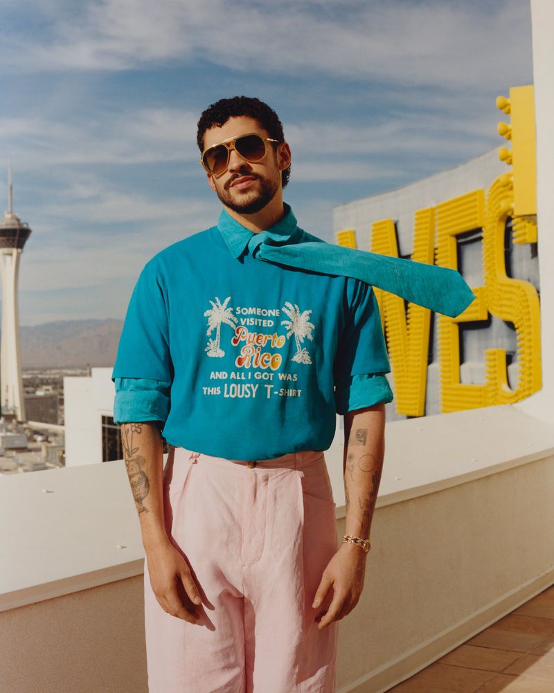 Bad Bunny wearing aviator sunglasses, a blue t-shirt, and pink trousers on a rooftop with a tall tower in the background.