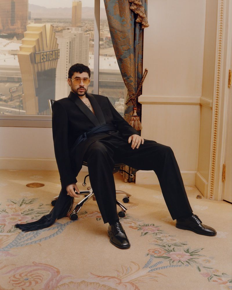 Bad Bunny seated in a rolling office chair wearing a black suit and tinted sunglasses inside a hotel room.