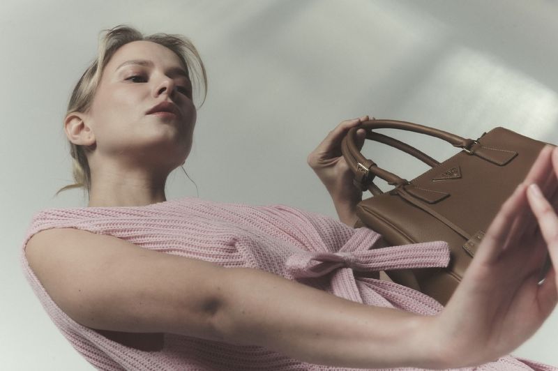 Low-angle fashion portrait of a model in a pink knit top holding a brown leather handbag against a plain background