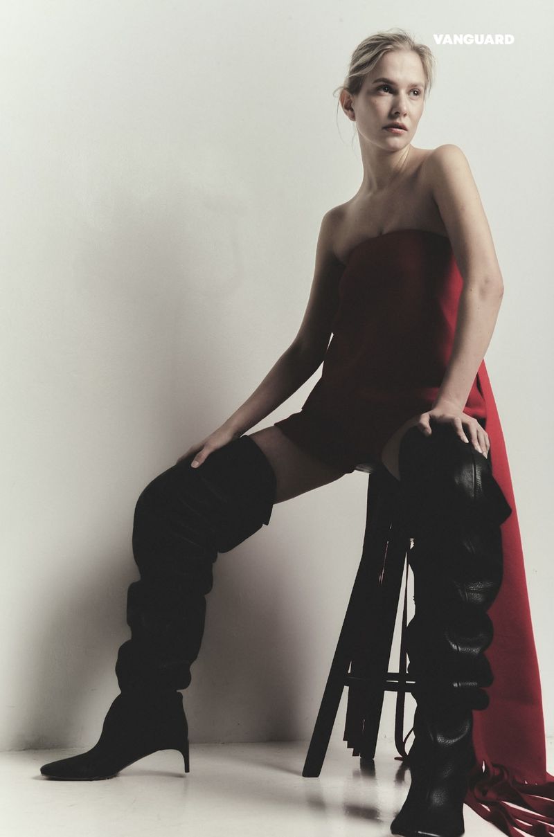 Model seated on stool wearing a red strapless mini dress and tall black boots against a plain white background