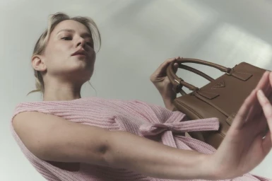 Low-angle fashion portrait of a model in a pink knit top holding a brown leather handbag against a plain background