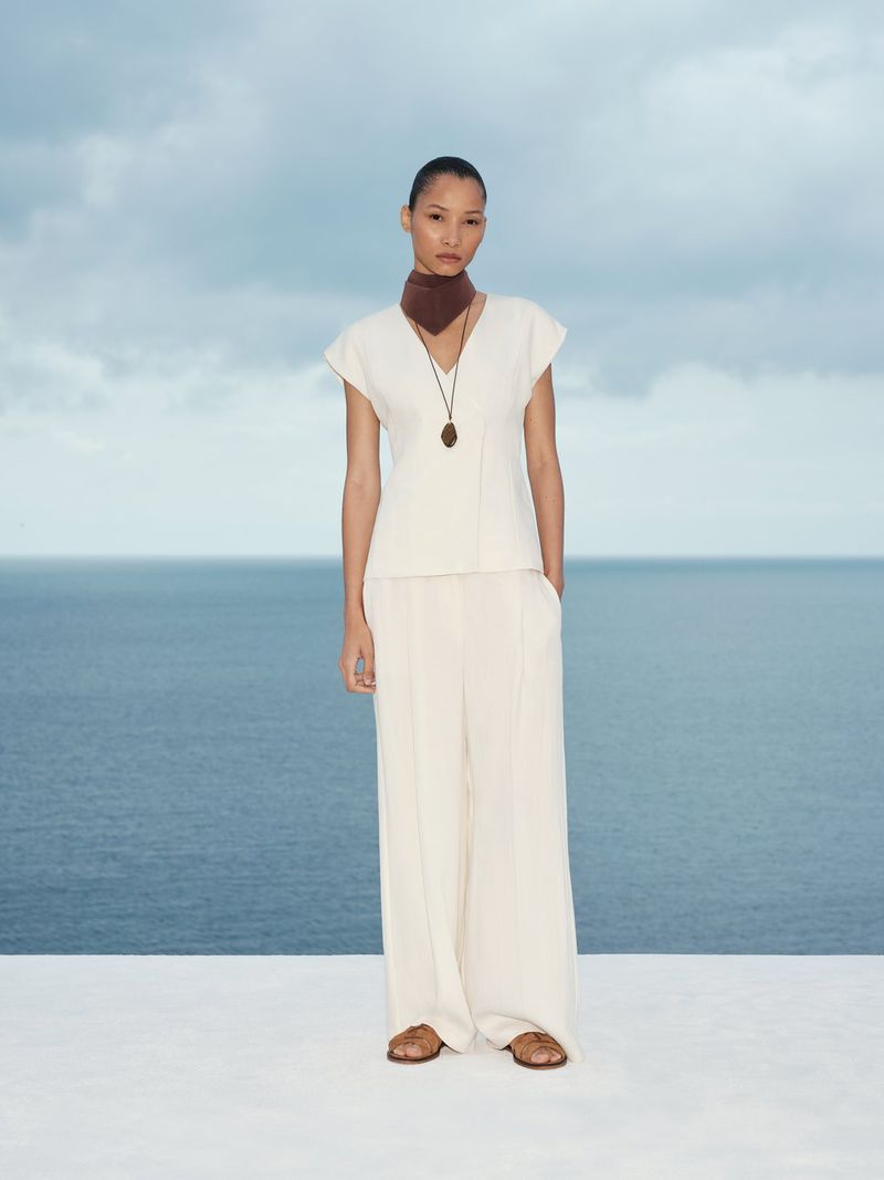 Model wearing a white top and trousers with a brown neck scarf standing against an ocean view backdrop.