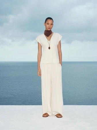 Model wearing a white top and trousers with a brown neck scarf standing against an ocean view backdrop.