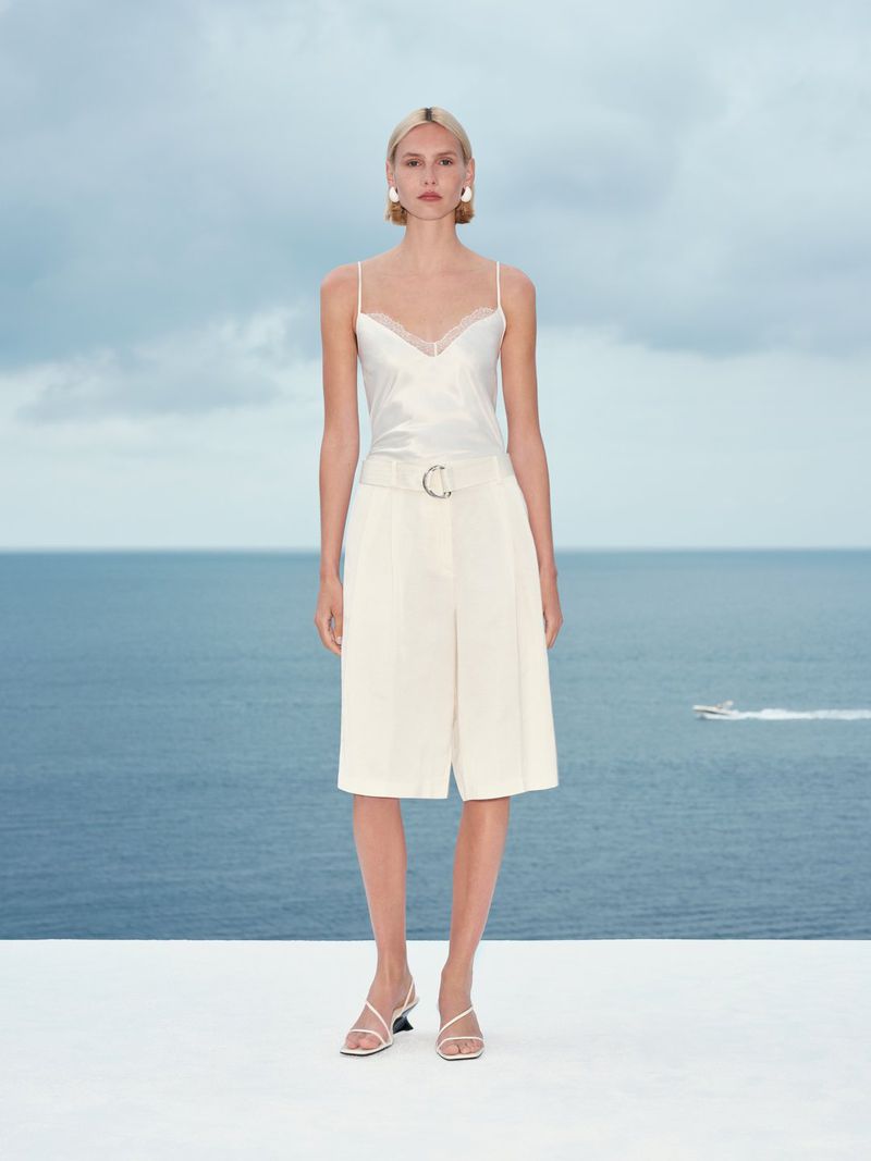 Model standing on a white terrace wearing a white camisole and cream tailored shorts against an ocean view