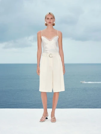 Model standing on a white terrace wearing a white camisole and cream tailored shorts against an ocean view