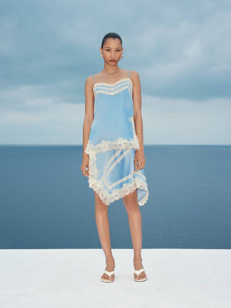 Model wearing a light blue slip dress with white lace trim standing on a platform against an ocean backdrop.