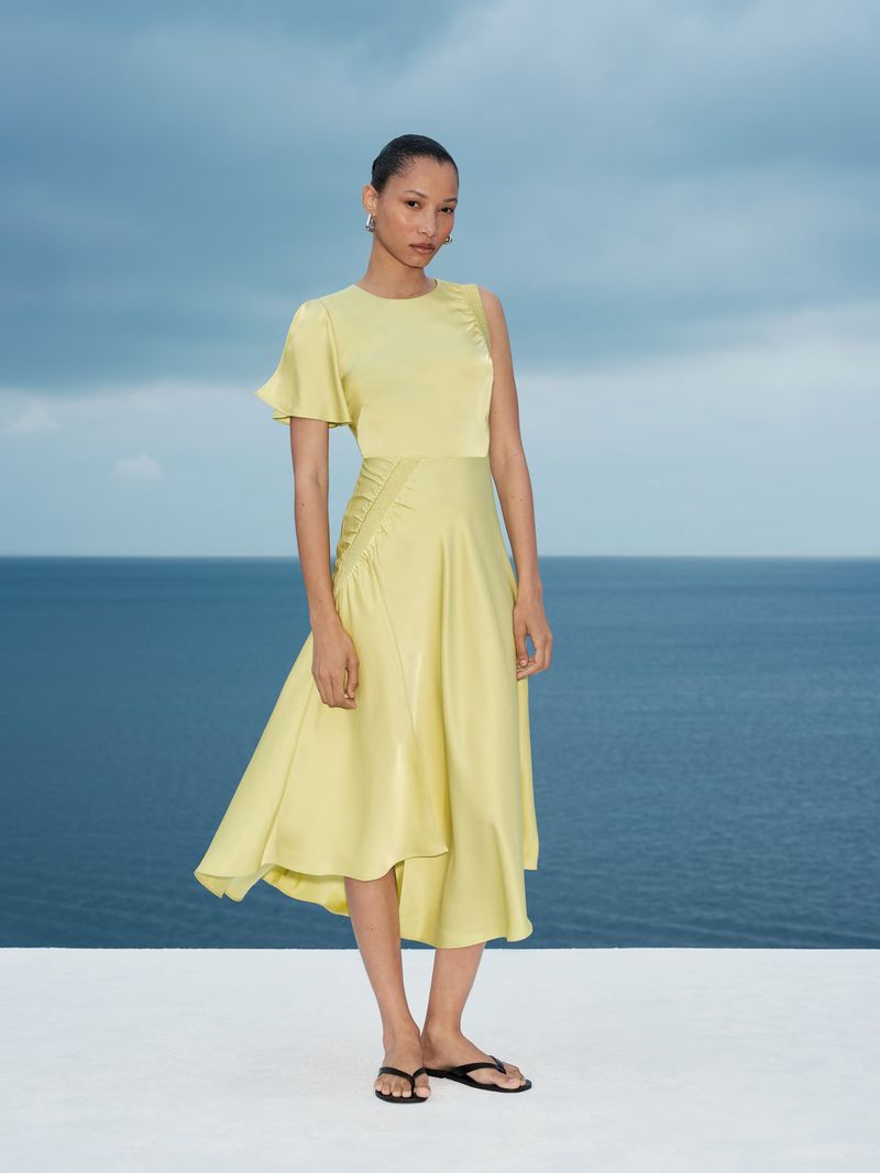 Model standing on a white surface wearing a yellow midi dress against a blue ocean and cloudy sky background